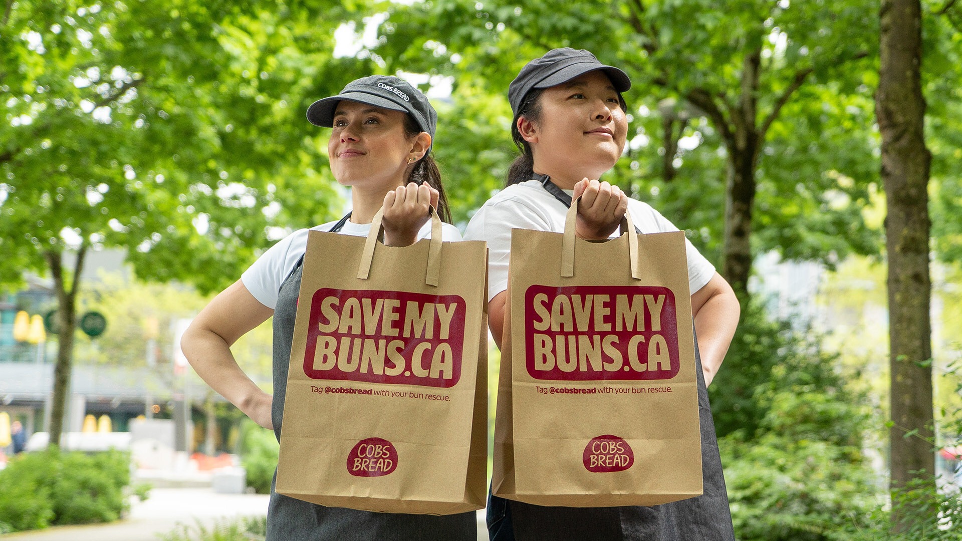 | PR In Canada Two women in caps hold brown paper bags with the'SAVEMY BUNS.CA' logo, posing in a sunny green park (volunteer-style fundraiser).