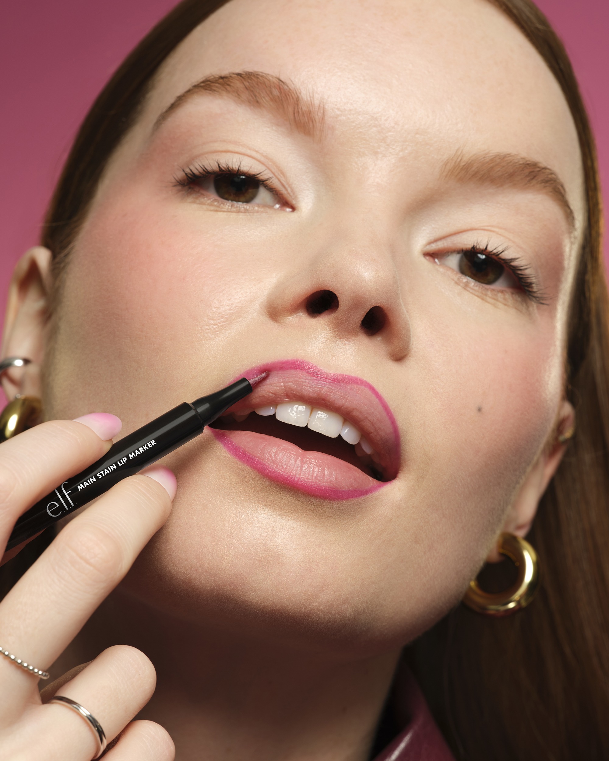 Close-up of a woman's lips being lined with pink lip liner by a makeup artist, showing detail of lipstick and eye makeup against a pink background.