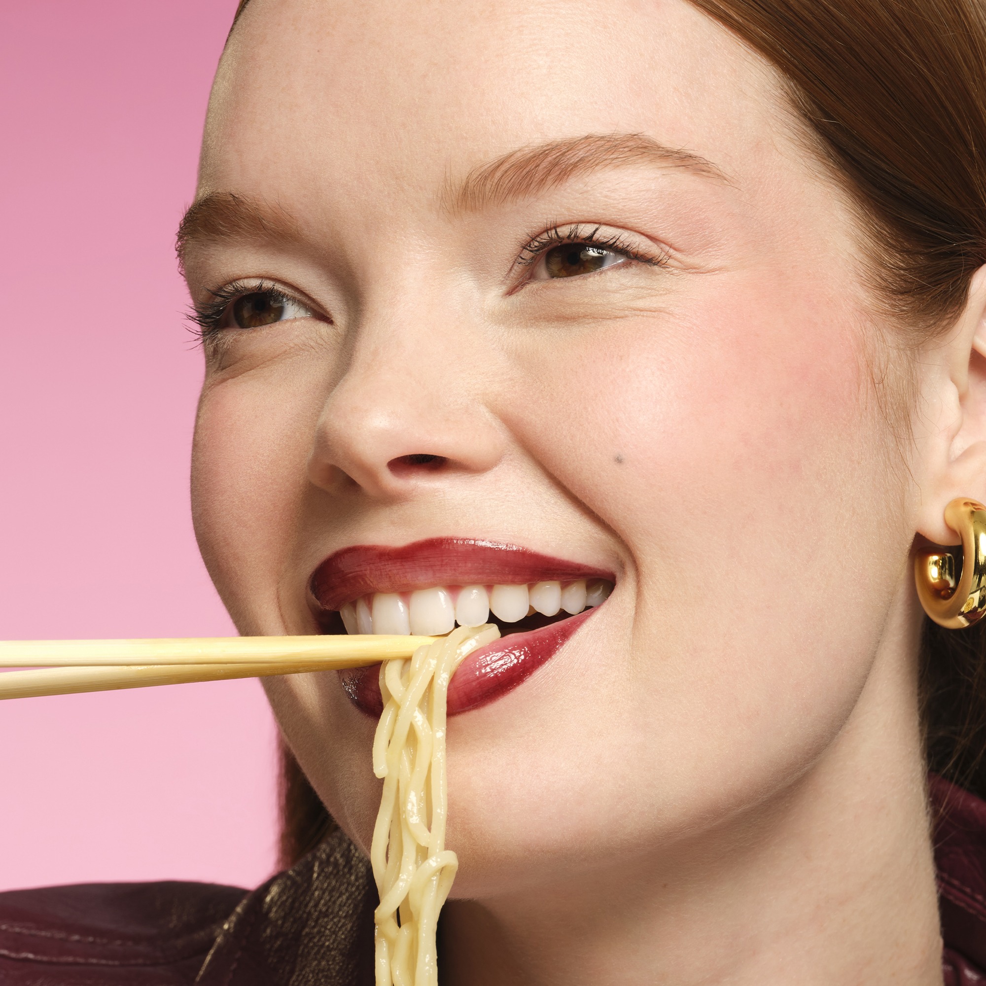 Close-up of a smiling woman's mouth with noodles held between chopsticks near her lips against a pink background.