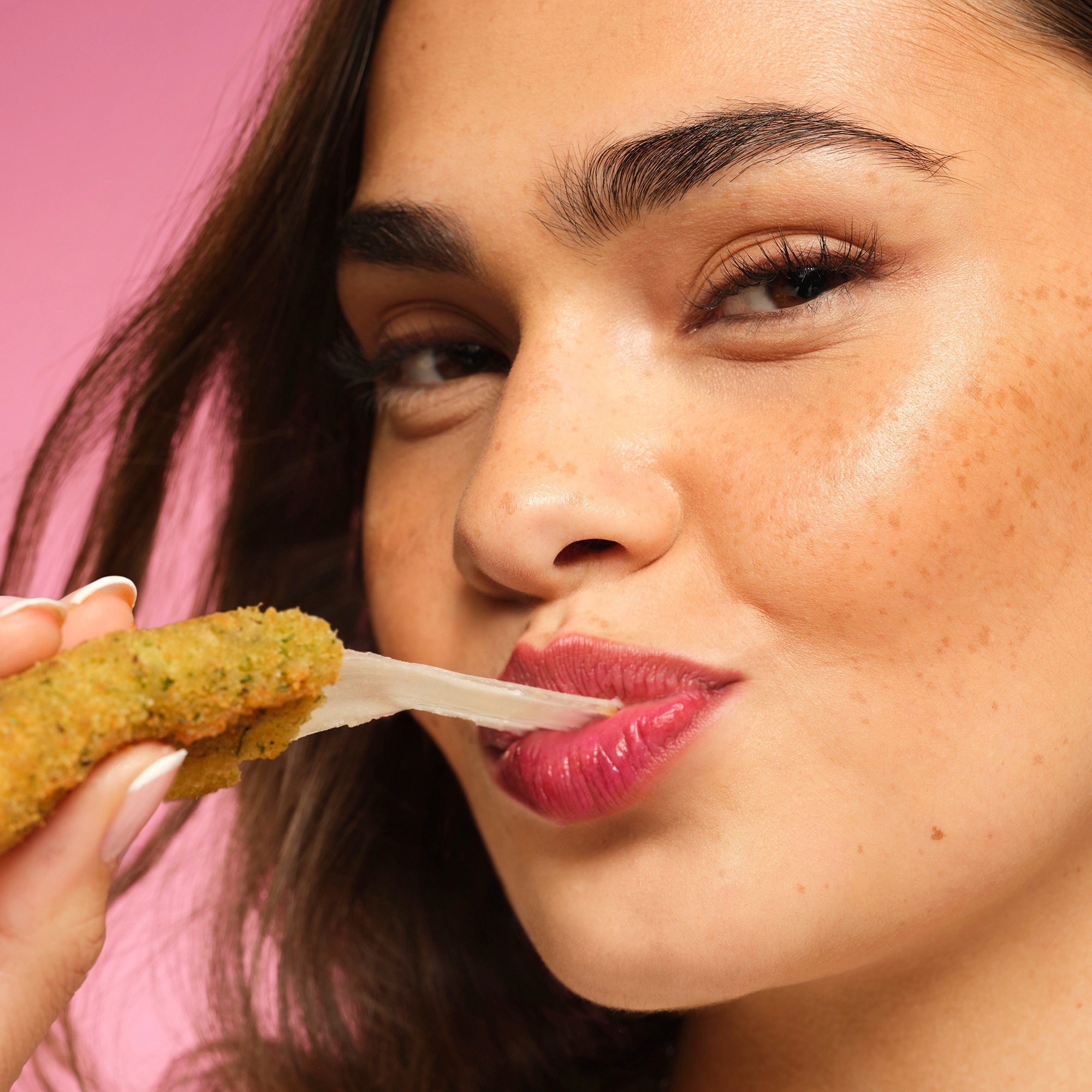 Close-up of a woman about to bite a cracker held near her lips, with pink background and visible eye and eyebrow details.