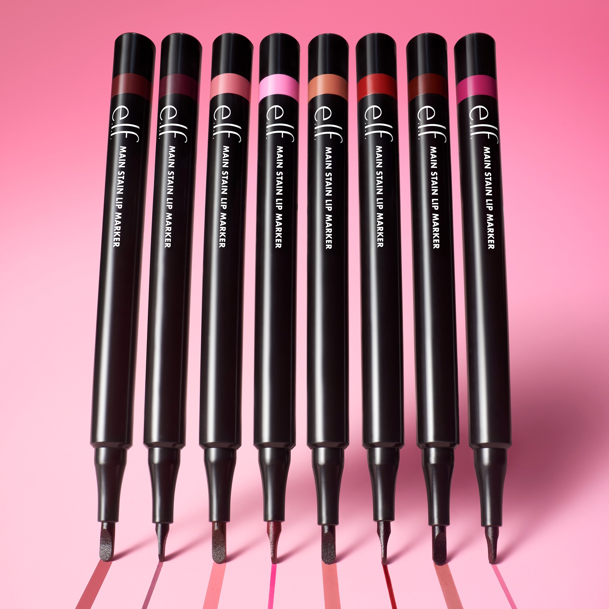 Row of black lip products with color swatches on a pink background, showing shades from pink to red (lipsticks or lip liners).